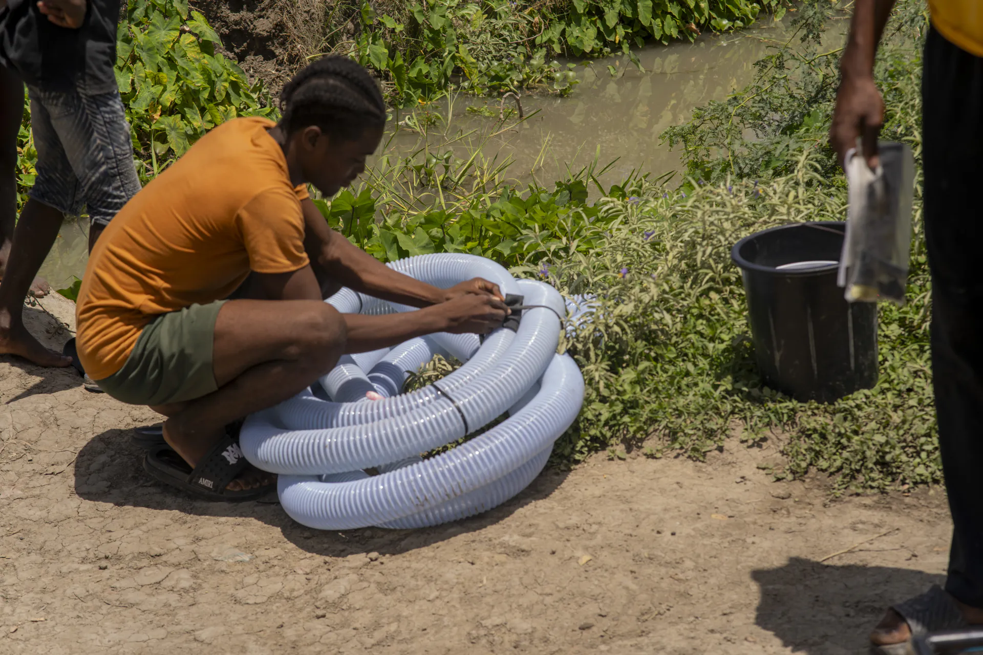 Un membre de la communauté, accroupi au bord d'un canal d'irrigation à Aux-Sources, raccorde un rouleau de tuyau d'irrigation transparent.